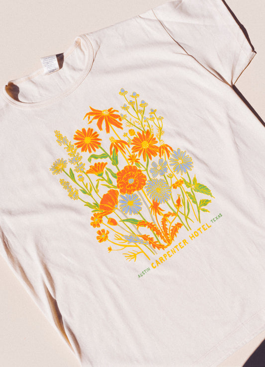 Photograph of beige cotton T-shirt with colorful floral print with text below it that reads 'CARPENTER HOTEL AUSTIN TEXAS' laying on top of beige background lit by natural sunlight
