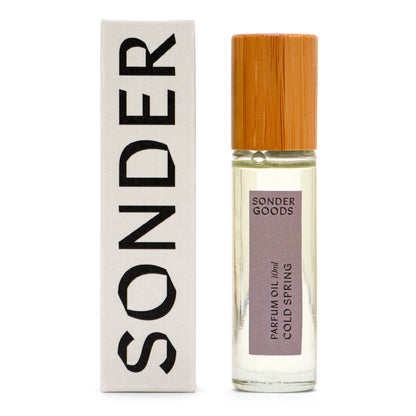 A small, clear glass bottle with a wooden cap, labeled 'Sonder Goods Parfum Oil Cold Spring', next to its white box packaging with the word 'SONDER' in black text.